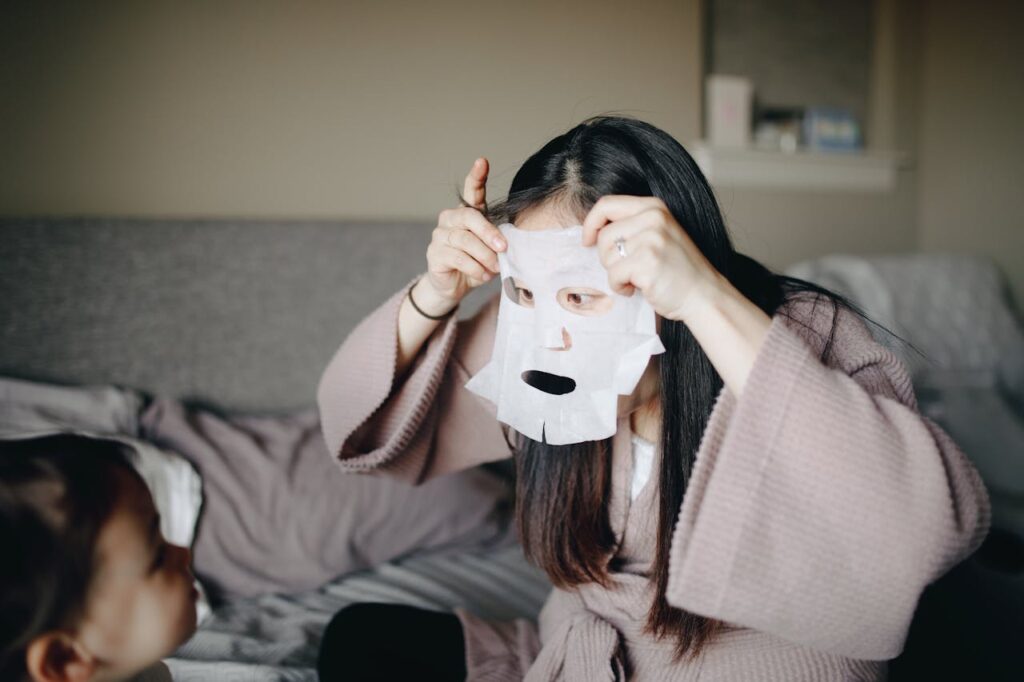 A mum enjoying a self-care moment with a facial mask while bonding with her baby. Skincare for mums in Malaysia is just as important as baby skincare.