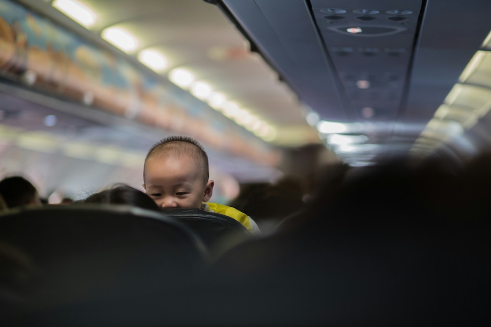 Baby on Airplane Looking Over Seat – A curious baby peeks over an airplane seat during a flight, highlighting the importance of travel essentials to keep babies comfortable in new environments.