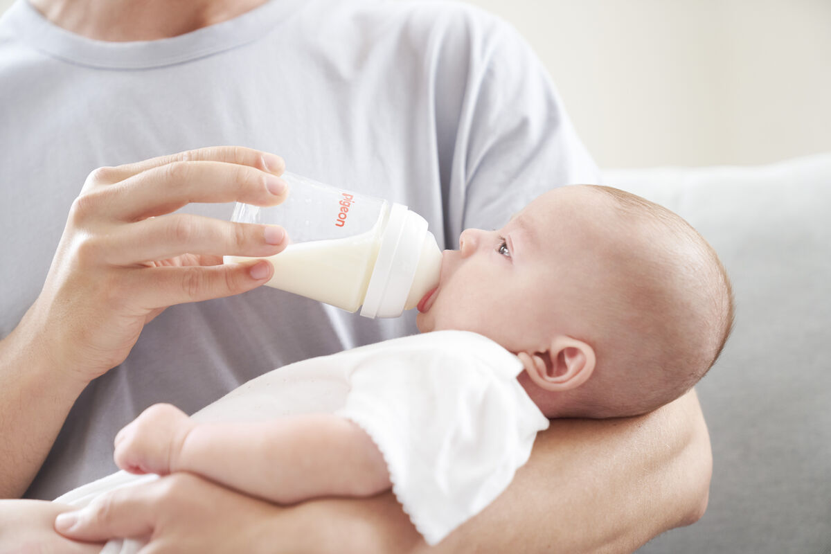 A father holds his baby, who is calmly drinking milk from a white Pigeon SofTouch bottle.
