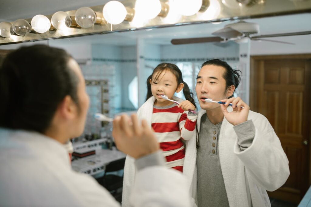 Father and daughter brushing teeth together in bathroom mirror during family oral care routine.