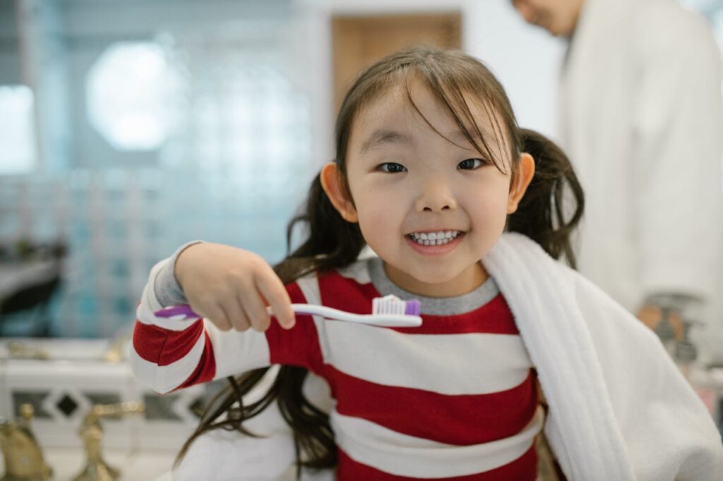 Smiling preschool girl brushing teeth in bathroom with toothbrush and towel over shoulders.