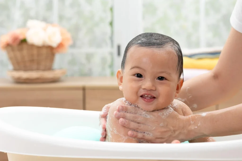 Smiling newborn baby being gently bathed by a parent in a baby bathtub with warm water and soft bubbles.